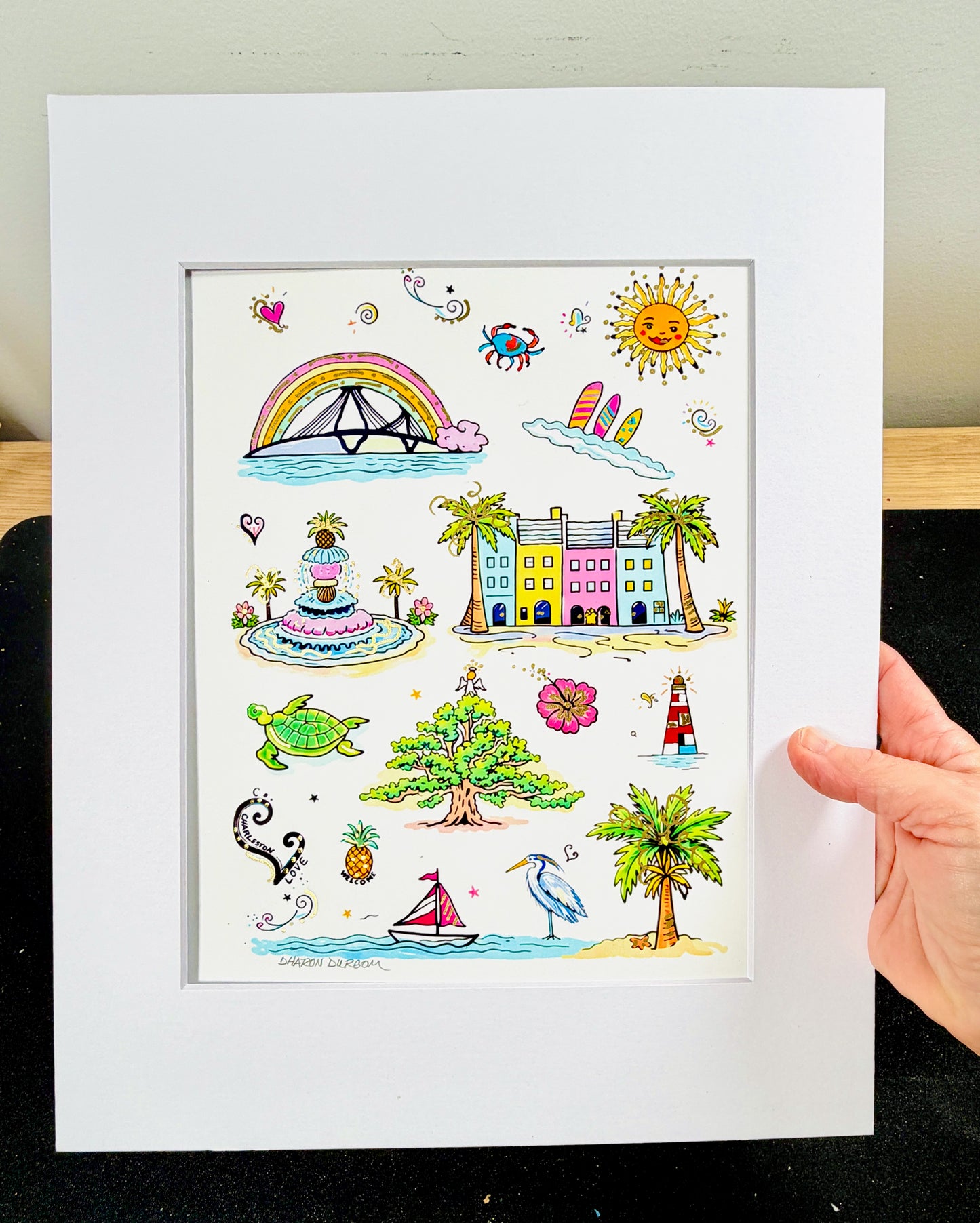 Charleston Charm Artist Embellished Print flat lay showing all Charleston icons including Rainbow Row, Ravenel Bridge, Pineapple Fountain, Angel Oak, and Sullivan's Island Lighthouse with gold accents